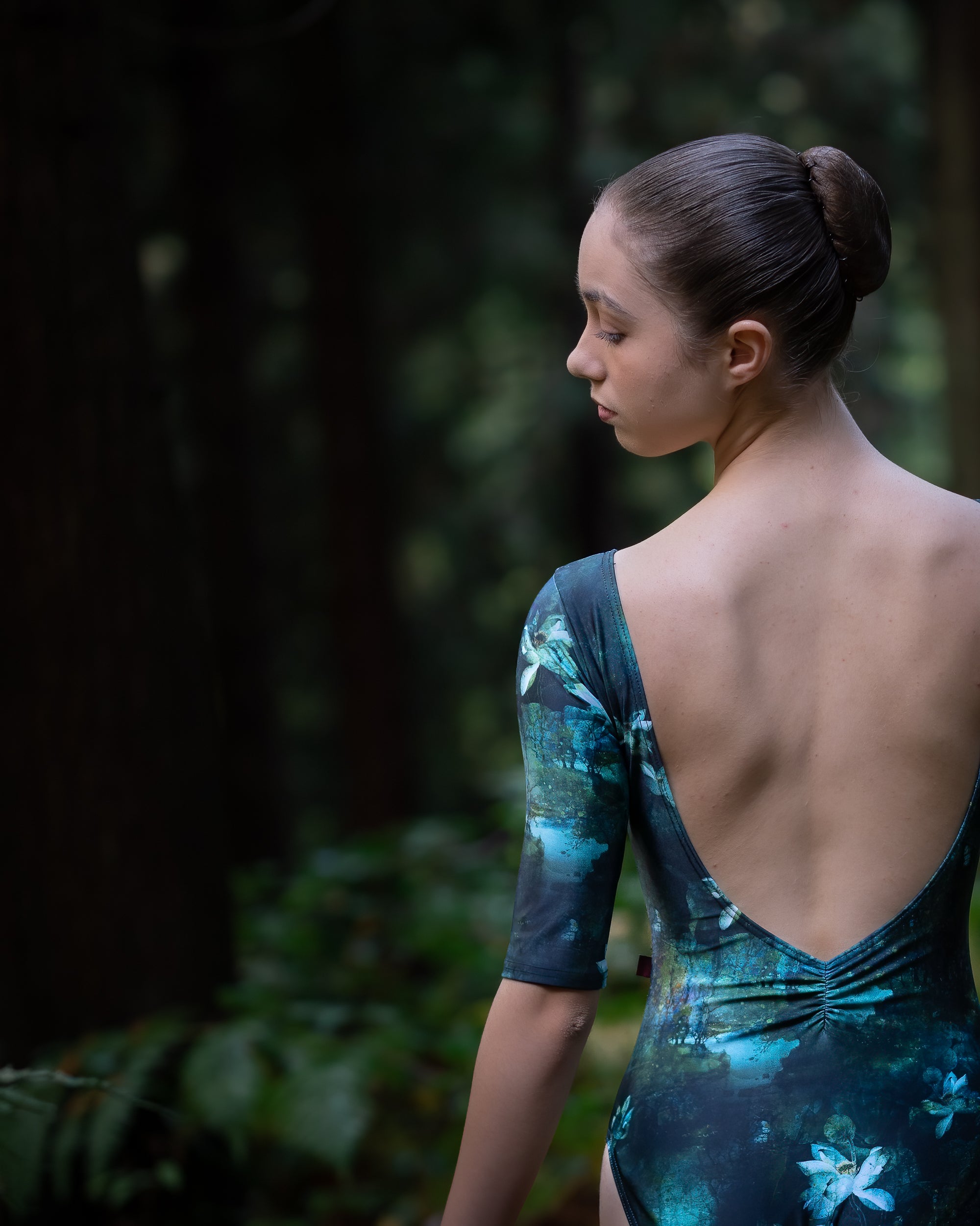 Dancer wearing green forest print girls dance leotard with sleeves in woodland setting. Showing low back leotard.