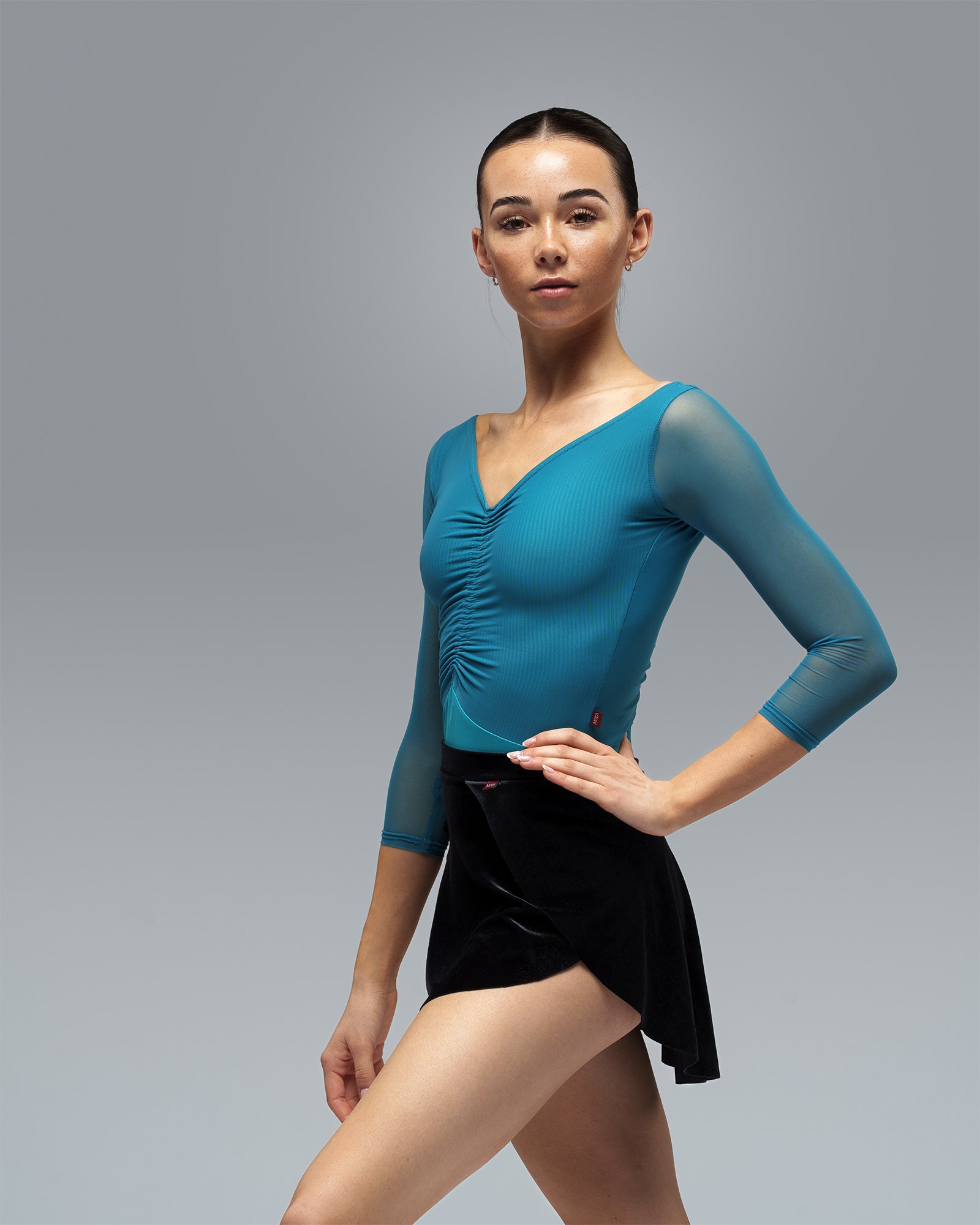 Dancer wearing teal green blue leotard with mesh sleeves and central ruche in ballet pose with black SAb ballet skirt.