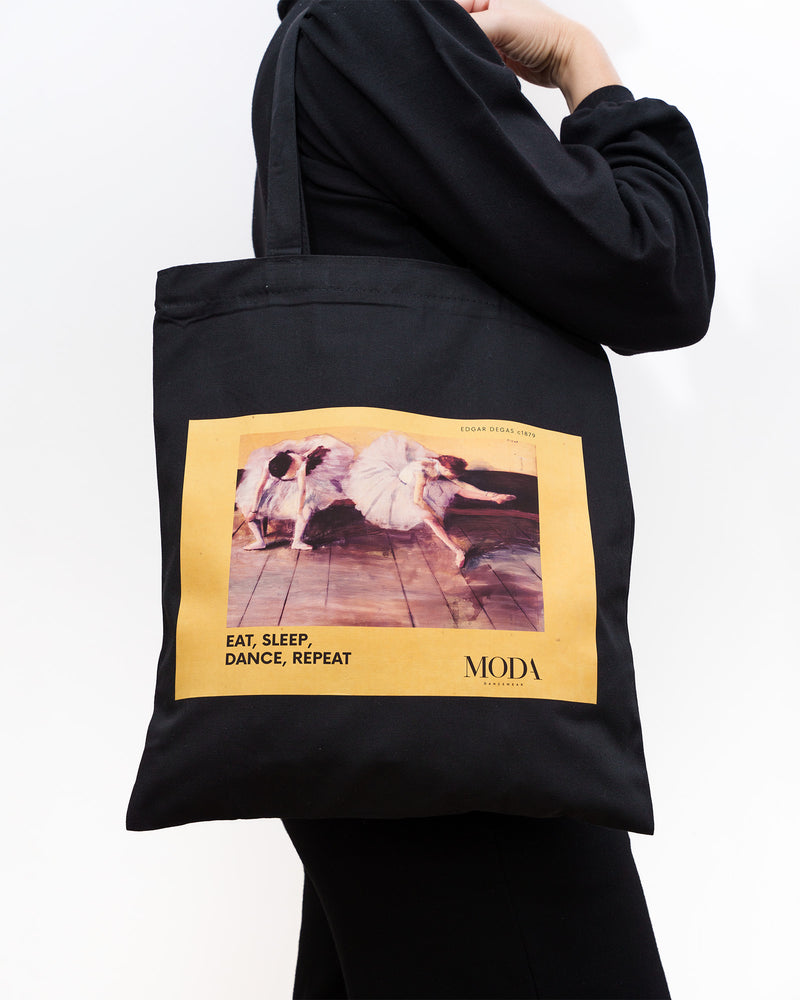 Black Edgar Degas ballerina dancer tote bag with a yellow graphic and text held by a person wearing a black hoodie.